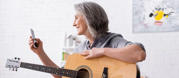 Sorrindo mulher madura com cabelos grisalhos segurando smartphone enquanto aprende a tocar guitarra acústica, banner — Fotografia de Stock