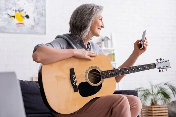 Sonriente mujer madura con pelo gris sosteniendo teléfono inteligente mientras aprende a tocar la guitarra acústica - foto de stock