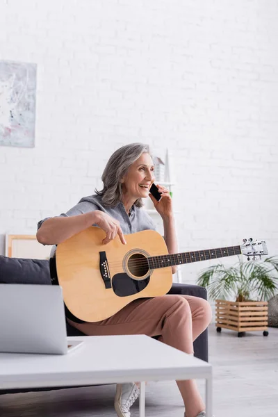 Smiling mature woman with grey hair talking on smartphone while holding acoustic guitar — Stock Photo