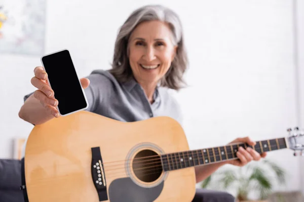 Mulher madura borrada e sorridente com cabelo grisalho segurando smartphone com tela em branco e guitarra acústica — Fotografia de Stock