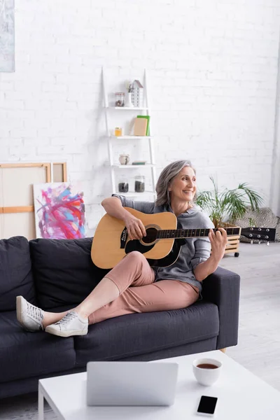 Glückliche Frau mittleren Alters spielt Akustikgitarre in der Nähe von Gadgets und einer Tasse Tee auf dem Couchtisch — Stockfoto