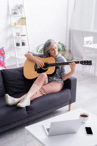 Frau mittleren Alters spielt Akustikgitarre in der Nähe von Geräten und einer Tasse Tee auf einem Couchtisch — Stockfoto
