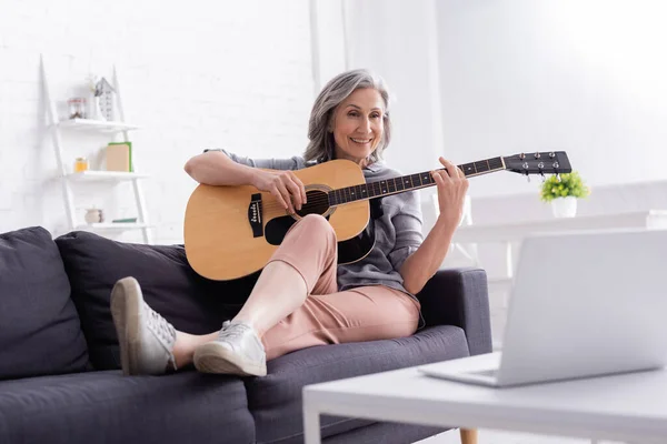 Alegre mujer de mediana edad tocando la guitarra acústica en el sofá cerca del ordenador portátil en la mesa de café - foto de stock