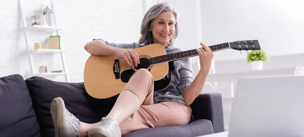 Весела жінка середнього віку грає на акустичній гітарі на дивані біля ноутбука, банер — стокове фото
