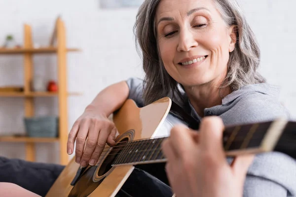 Mujer madura positiva con pelo gris tocando la guitarra acústica en primer plano borroso - foto de stock