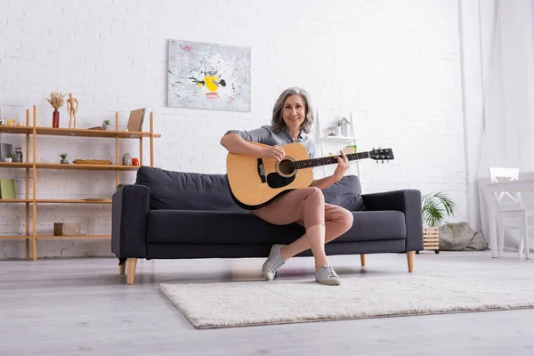 Mujer madura alegre con el pelo gris sentado en el sofá y tocando la guitarra acústica en la sala de estar - foto de stock