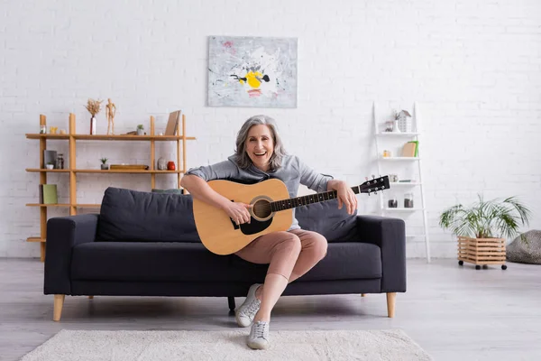 Mujer madura alegre con pelo gris sentado en el sofá con guitarra acústica - foto de stock