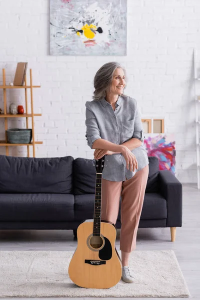 Довольная зрелая женщина с седыми волосами, стоящая с акустической гитарой возле дивана в гостиной — стоковое фото