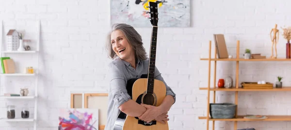 Mujer madura alegre con pelo gris de pie con guitarra acústica en la sala de estar, pancarta - foto de stock