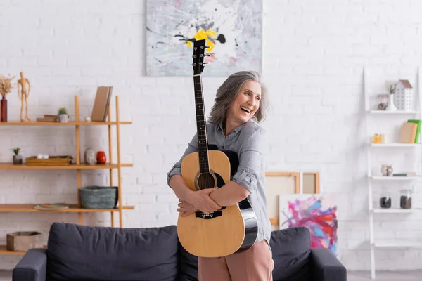 Fröhliche reife Frau mit grauen Haaren umarmt Akustikgitarre neben Couch im Wohnzimmer — Stockfoto