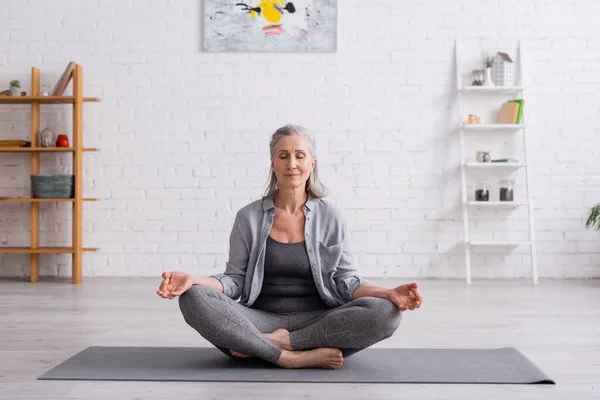 Mujer madura con pelo gris sentada en pose de loto en esterilla de yoga - foto de stock