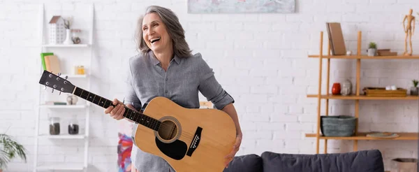 Mujer madura feliz con pelo gris de pie con guitarra acústica en la sala de estar, pancarta - foto de stock
