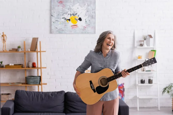 Mujer madura feliz con el pelo gris de pie con guitarra acústica y riendo en la sala de estar - foto de stock