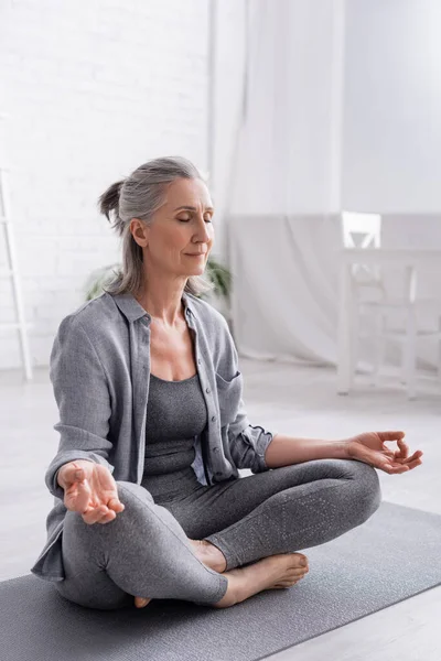 Reife Frau mit grauen Haaren sitzt in Lotus-Pose und meditiert auf Yogamatte — Stockfoto