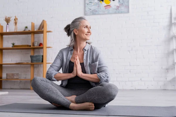 Щаслива зріла жінка з сірим волоссям, сидячи з молитовними руками в позі лотоса на йога килимок — стокове фото