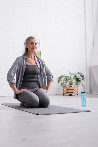 Mujer madura feliz con pelo gris practicando yoga cerca de la botella deportiva - foto de stock