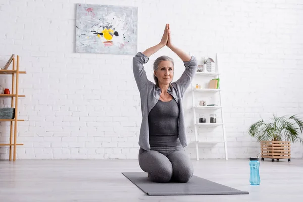 Femme mûre avec les cheveux gris et les mains priantes au-dessus de la tête pratiquant le yoga près de bouteille de sport — Photo de stock