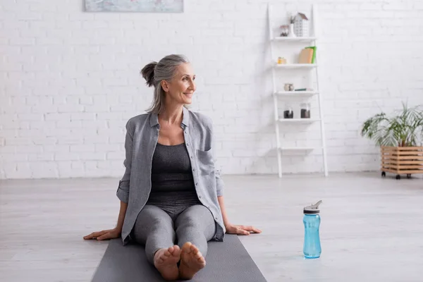 Mujer madura feliz con el pelo gris sentado en la estera del yoga cerca de la botella de deportes - foto de stock