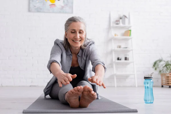 Mujer madura feliz con el pelo gris que estira en la estera del yoga cerca de la botella de los deportes - foto de stock