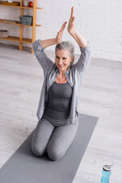 Mature woman with praying hands above head practicing yoga near sports bottle — Stock Photo