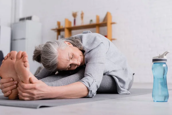 Middle aged woman with grey hair stretching on yoga mat near sports bottle — Stock Photo