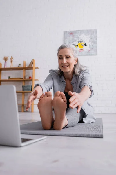 Femme mûre souriante avec les cheveux gris s'étendant sur le tapis de yoga près de l'ordinateur portable sur le premier plan flou — Photo de stock
