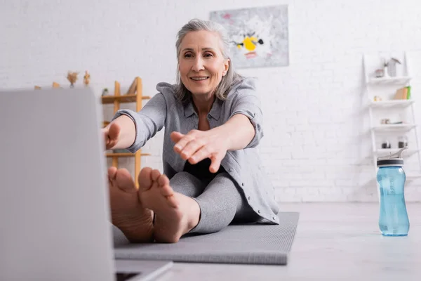 Mujer madura con pelo gris estirándose sobre esterilla de yoga cerca de botella deportiva - foto de stock