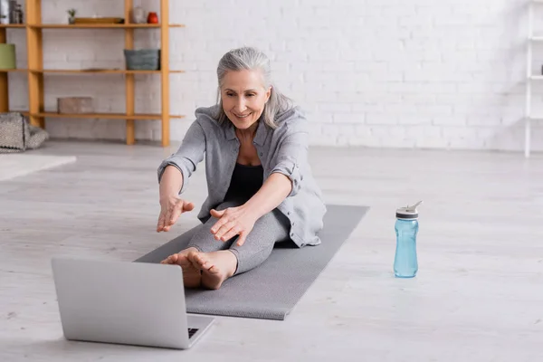 Femme mûre avec les cheveux gris étirant sur le tapis de yoga près de bouteille de sport et ordinateur portable — Photo de stock