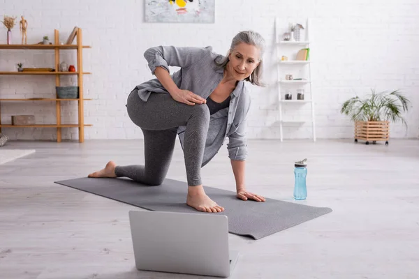 Mujer madura con el pelo gris practicando en la estera del yoga cerca de la botella de los deportes mientras que mira tutorial en el ordenador portátil - foto de stock