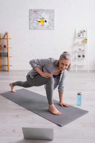 Mujer madura feliz con pelo gris practicando yoga en la estera cerca de la botella de deportes mientras ve el tutorial en el ordenador portátil - foto de stock