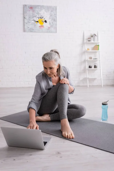 Mature woman with grey hair sitting on yoga mat while watching tutorial on laptop — Stock Photo