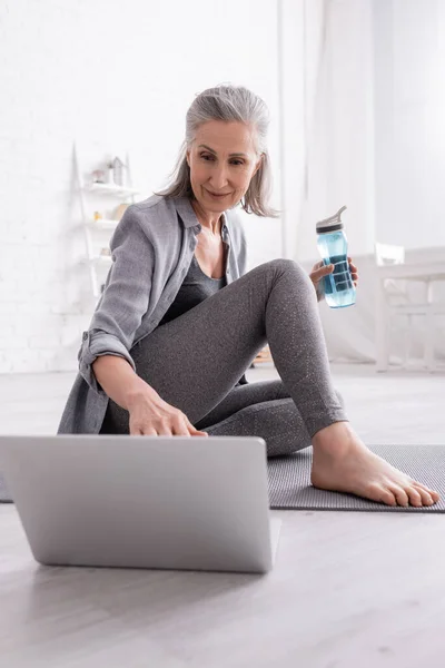 Mature woman with grey hair sitting on yoga mat while watching tutorial on laptop and holding sports bottle — Stock Photo