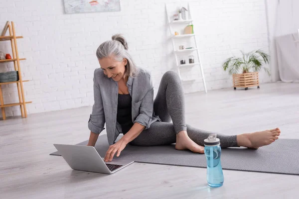 Mujer madura sonriente sentada en la esterilla de yoga mientras mira el tutorial en el portátil - foto de stock