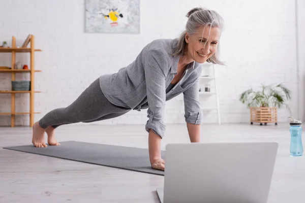 Glücklich reife Frau praktiziert und sieht Yoga-Tutorial auf verschwommenem Laptop — Stockfoto