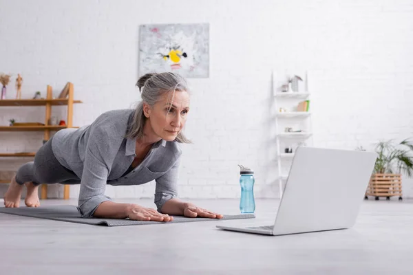 Femme d'âge moyen faisant planche sur tapis de fitness près d'un ordinateur portable — Photo de stock