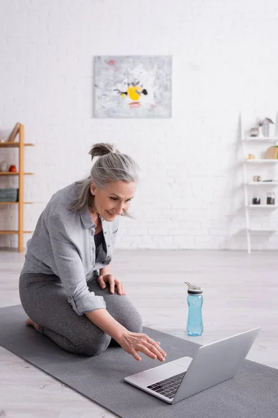 Souriant femme d'âge moyen assis sur tapis de fitness et à l'aide d'un ordinateur portable — Photo de stock