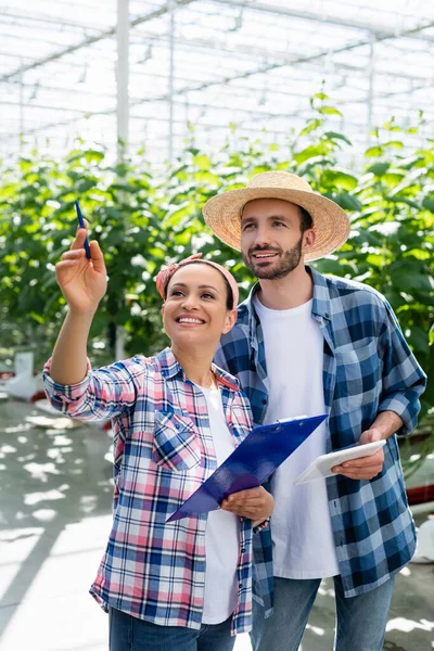 Smiling african american woman pointing with pen near farmer in greenhouse — Stock Photo