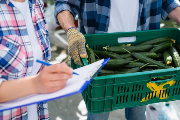 Partial view of farmer with box of fresh cucumbers near woman writing on clipboard on blurred foreground — Stock Photo