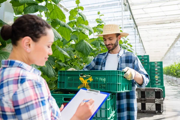 Farmer holding plastic box with harvest near african american colleague writing on clipboard on blurred foreground — Stock Photo