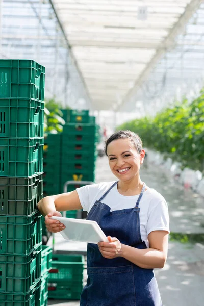 Alegre agricultor afroamericano de pie con tableta digital cerca de cajas de plástico en invernadero - foto de stock