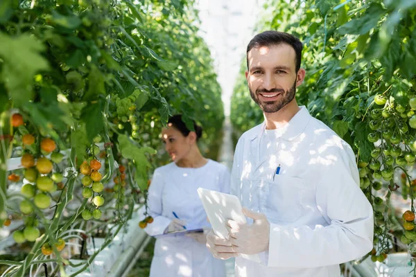 Lächelnder Qualitätsprüfer mit digitalem Tablet in der Nähe eines amerikanischen Kollegen, der auf verschwommenem Hintergrund arbeitet — Stockfoto