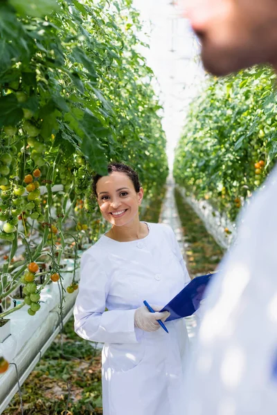 Africano americano calidad inspector con portapapeles sonriendo a cámara cerca colega en borrosa primer plano - foto de stock