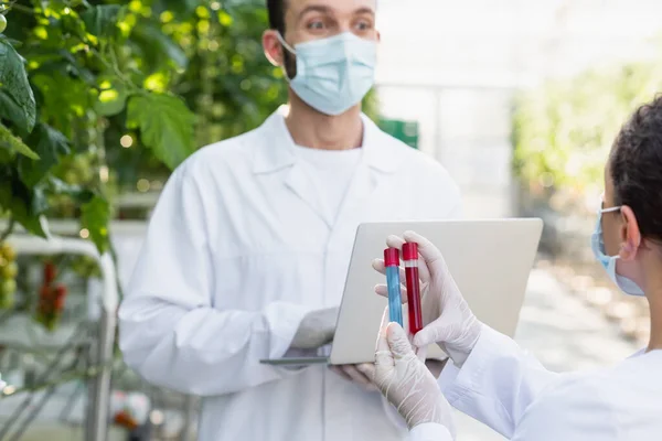 Qualitätsinspektor in medizinischer Maske mit Laptop in der Nähe eines afrikanischen amerikanischen Kollegen, verschwommener Hintergrund — Stockfoto