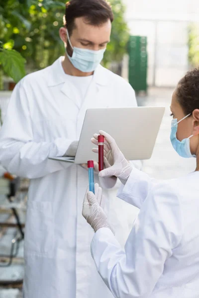 Inspetor de qualidade afro-americano em tubos de teste de retenção de máscara médica perto de colega com laptop — Fotografia de Stock