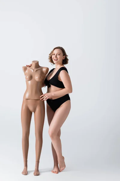 Full length of overweight and smiling young woman in swimsuit near plastic mannequin on white — Stock Photo