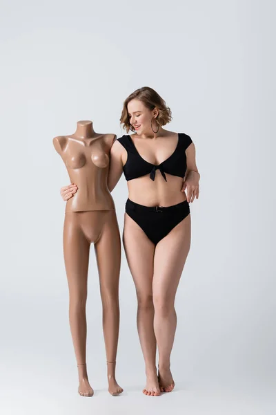 Full length of overweight and happy young woman in swimsuit hugging plastic mannequin on white — Stock Photo