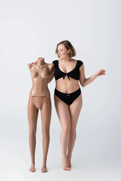 Volle Länge der übergewichtigen und lächelnden Frau im Badeanzug in der Nähe von Plastik-Schaufensterpuppe auf weiß — Stockfoto