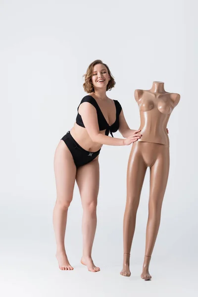 Full length of overweight and barefoot young woman laughing near plastic mannequin on white — Stock Photo