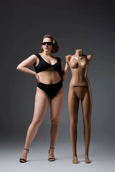 Übergewichtige junge Frau in Badeanzug und Sonnenbrille posiert mit Plastik-Schaufensterpuppe auf grau — Stockfoto