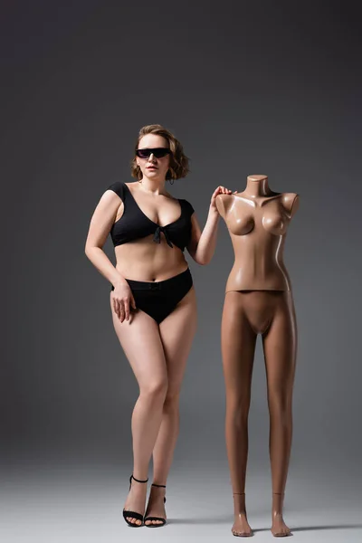 Übergewichtige junge Frau in Badeanzug und Sonnenbrille posiert in der Nähe von Plastik-Schaufensterpuppe auf grau — Stockfoto